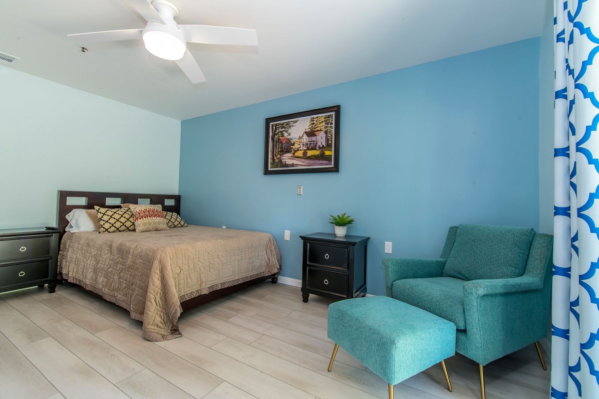 A cozy bedroom with a blue accent wall, featuring a bed, teal chair with matching ottoman, and decorative artwork.