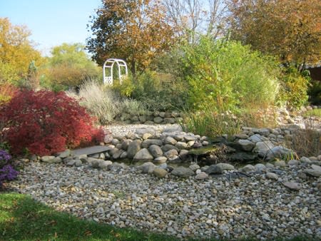 A serene garden landscape featuring a small pond surrounded by rocks and lush greenery, with trees displaying autumn foliage.