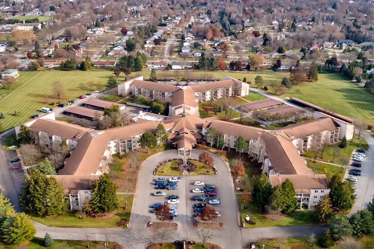 Aerial view of a large building complex situated in a suburban area with surrounding greenery and a parking lot in the foreground.