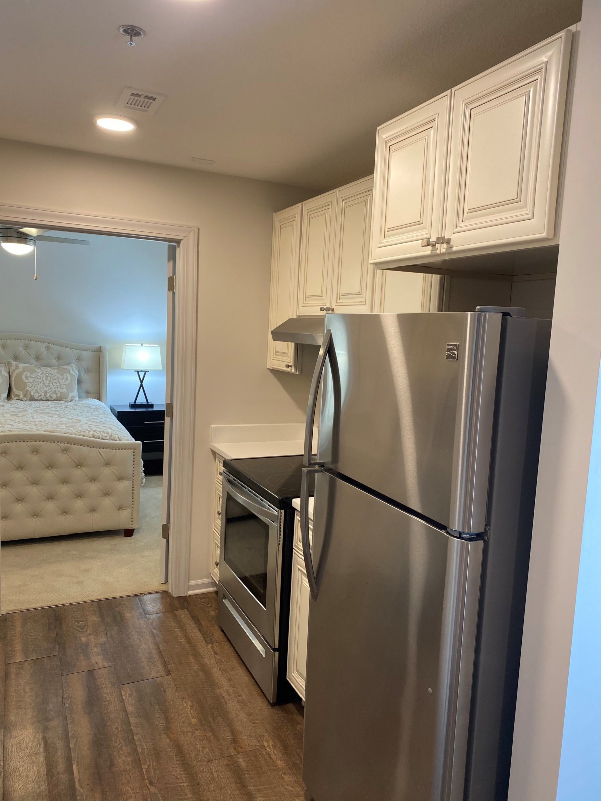 A view of a small modern kitchen with stainless steel appliances, leading into a bedroom.