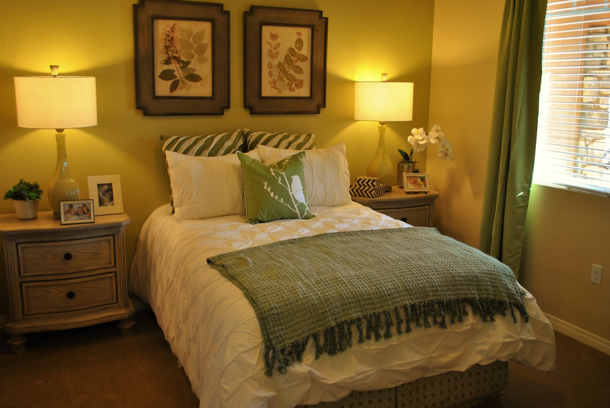 A cozy bedroom with a warm color scheme featuring framed wall art, a well-made bed with pillows and a throw blanket, and a bedside lamp providing soft lighting.
