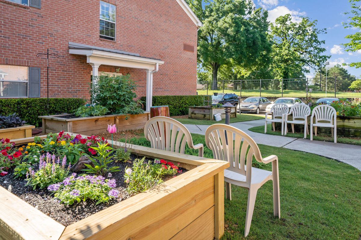 A cozy outdoor garden area with chairs and a flower bed next to a brick building on a sunny day.