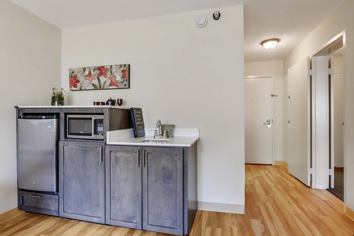 A small kitchenette area with a microwave on top of gray cabinets, adjacent to a hallway with wooden flooring.