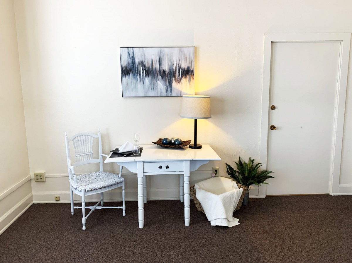 A minimalist room setup with a white table and chair, accompanied by a lamp, artwork, and plant.