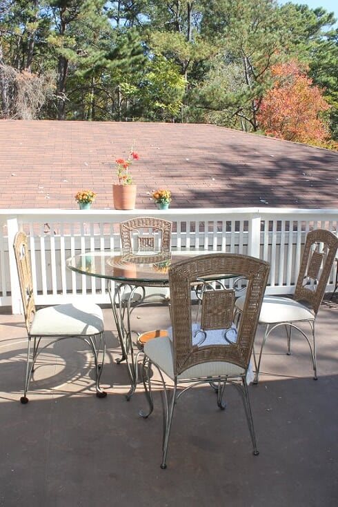 A sunny outdoor patio with a glass table and four chairs, decorated with potted plants in the background.