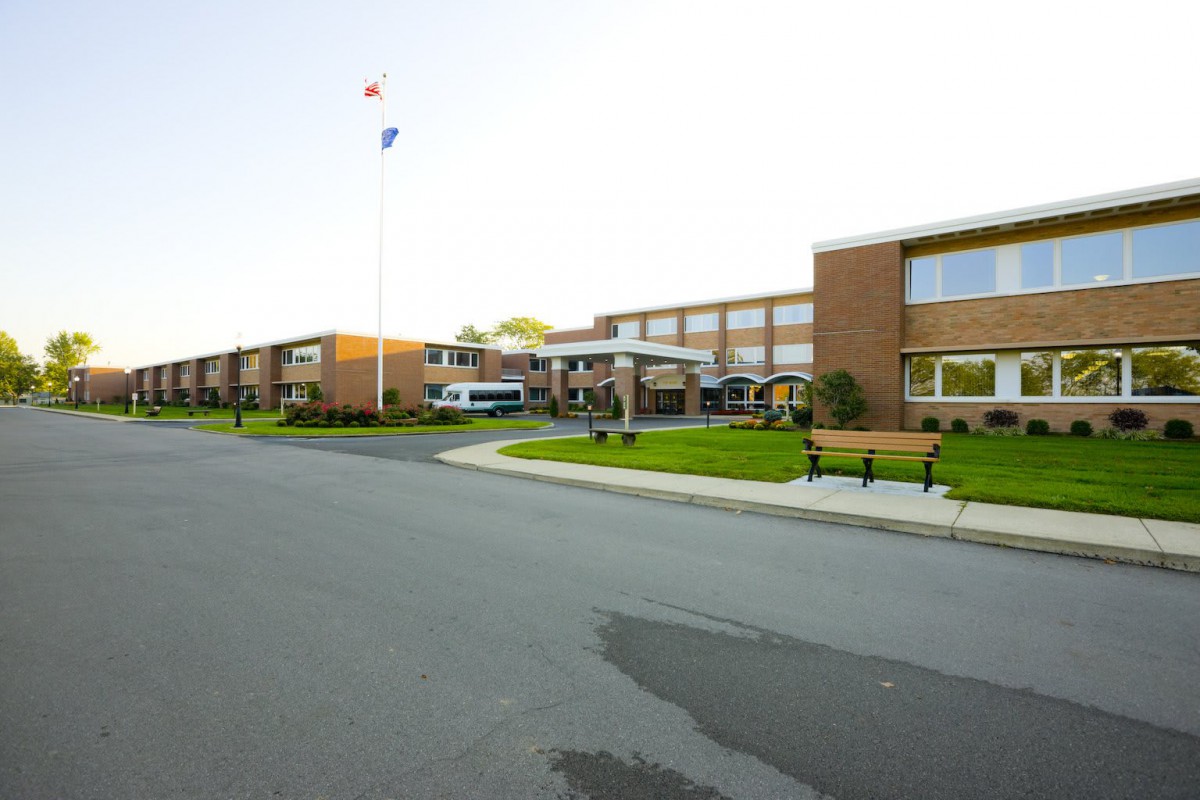 A brick school building with an American flag, benches, and adjacent road.