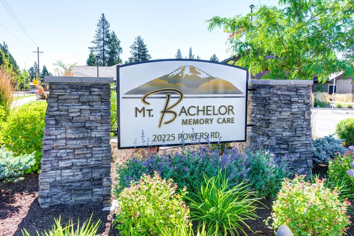 A decorative sign for Mt Bachelor Memory Care surrounded by greenery and a stone wall.