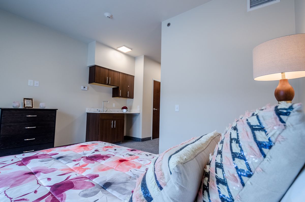 A modern bedroom featuring a floral bedspread and a small kitchenette in the background.