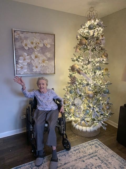 An elderly person in a wheelchair waves beside a beautifully decorated Christmas tree in a cozy living room setting.