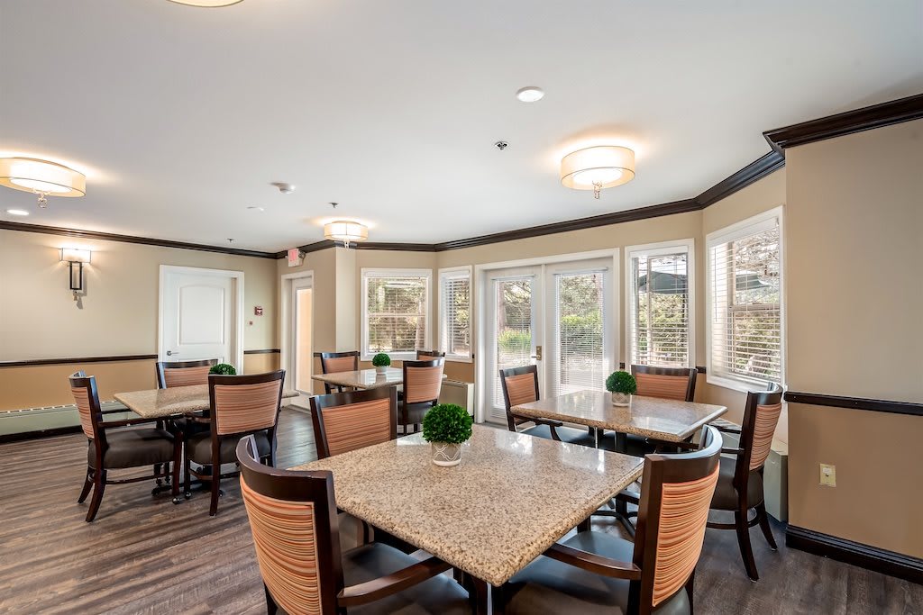A modern dining area with natural lighting, featuring tables and chairs arranged in a bright space with large windows.