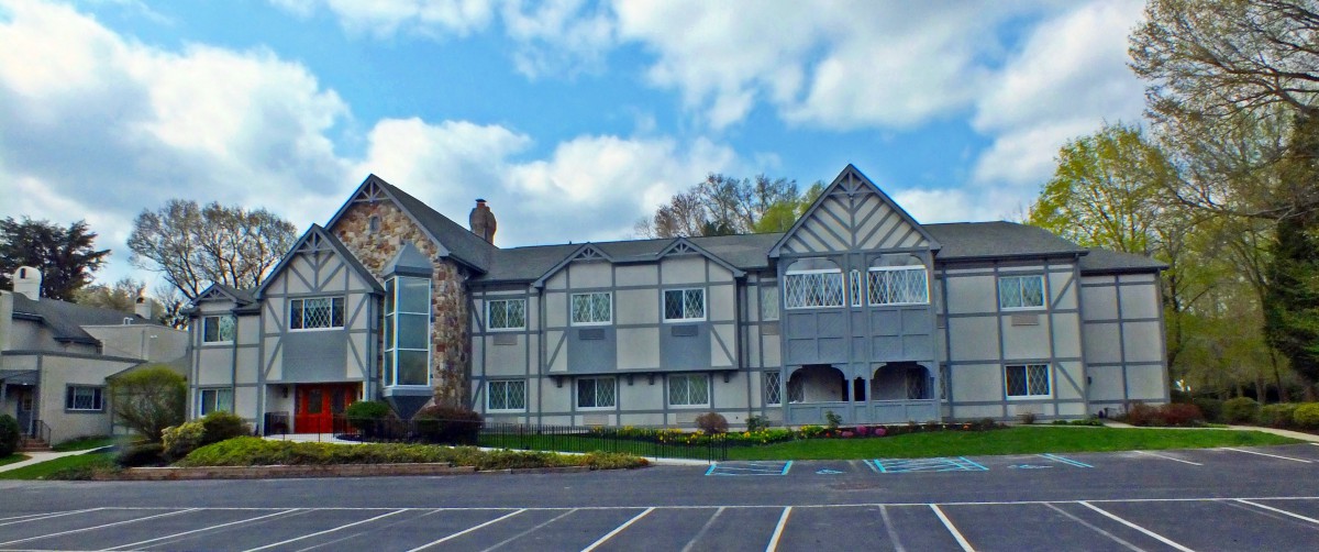 A Tudor-style building with a stone facade and gabled roof, set against a partly cloudy sky, is fronted by an empty parking lot.