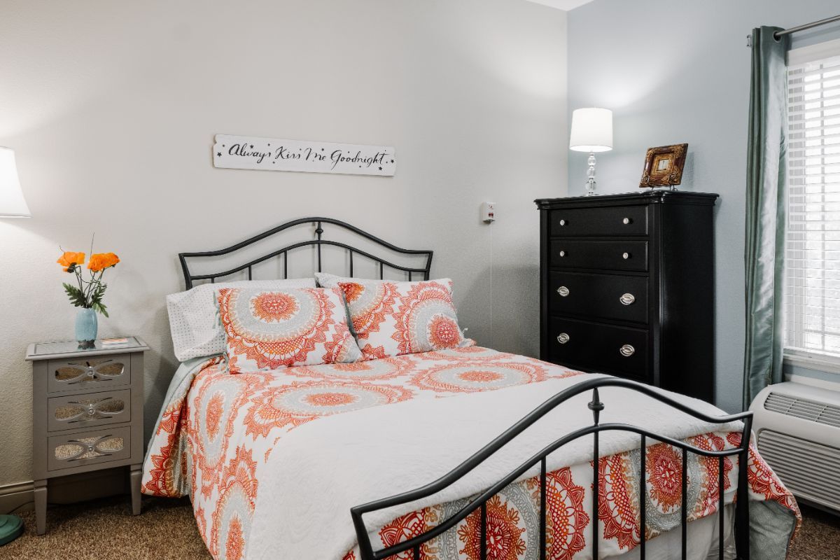 A cozy bedroom with patterned bedding and a black metal bedframe, accompanied by a dark wood dresser and a bedside lamp.