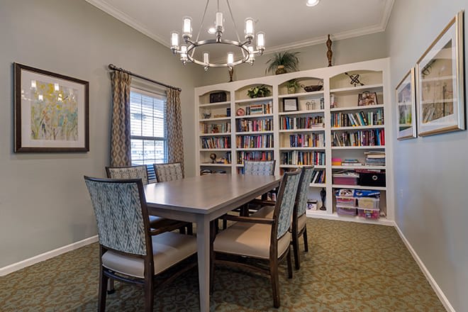A cozy dining room with a large table, surrounded by bookshelves and illuminated by a chandelier.