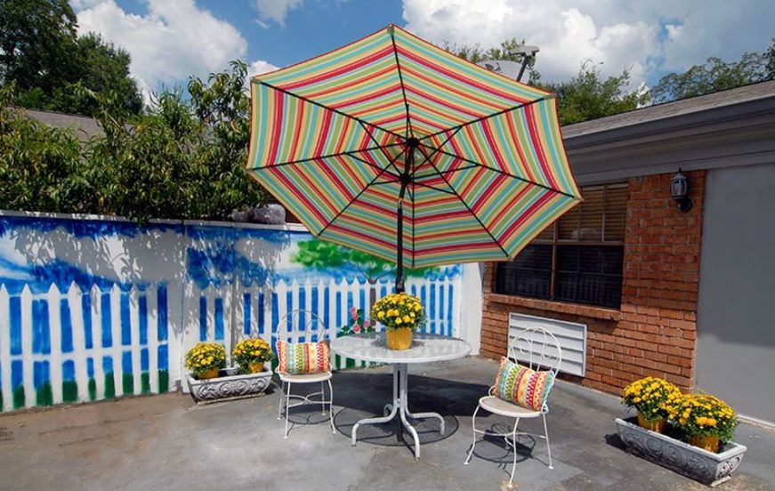 A cozy backyard patio with a colorful striped umbrella and a set of chairs around a table, adorned with potted flowers.