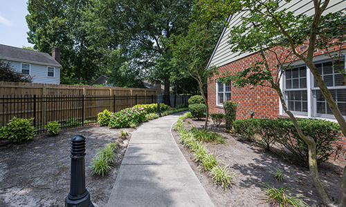 A serene garden pathway alongside a brick building with lush greenery and trees.