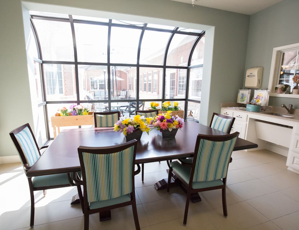 A bright dining room with a large window, decorated with colorful flowers on the wooden table.