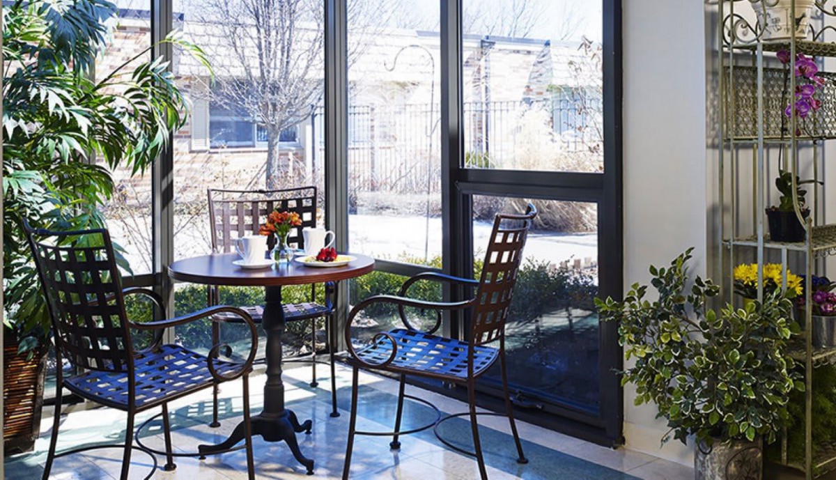 A sunlit room with a table and chairs by large windows, surrounded by plants.