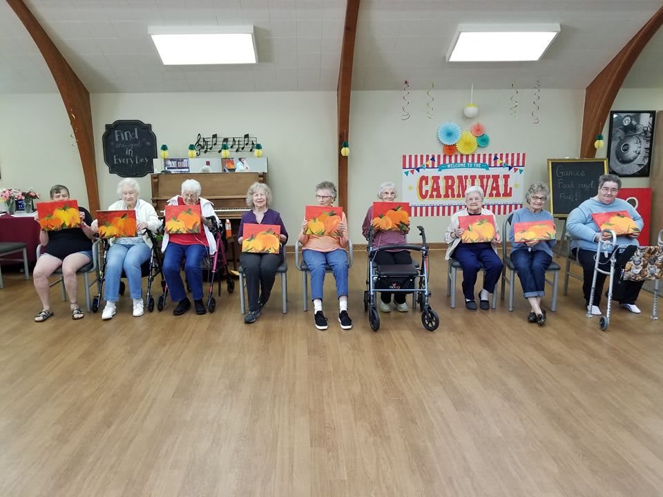 A group of seniors holding paintings seated indoors at a carnival-themed event.