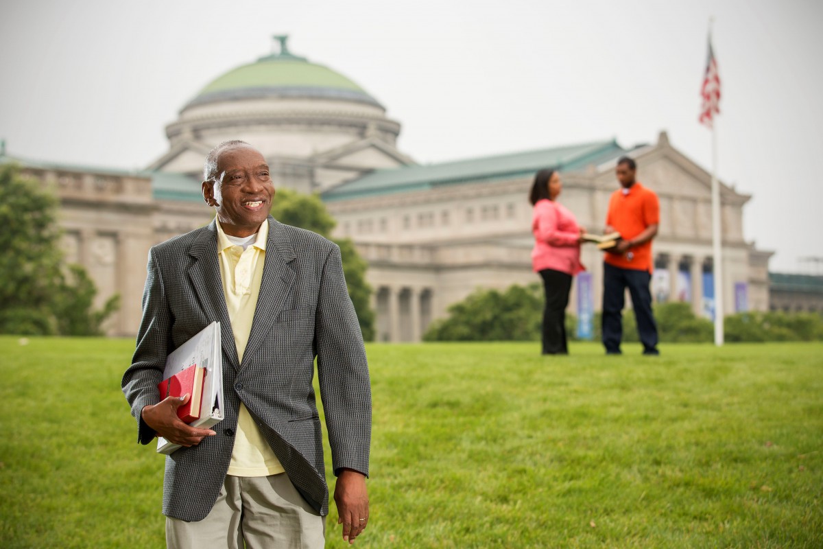A man holding books smiles while standing on a grassy area with a large building in the background.