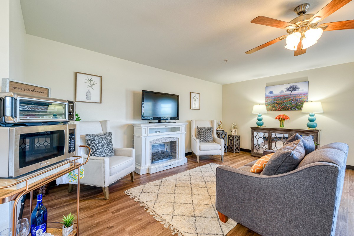A cozy living room with modern furniture, featuring a television above a fireplace, comfortable armchairs, and a sofa.