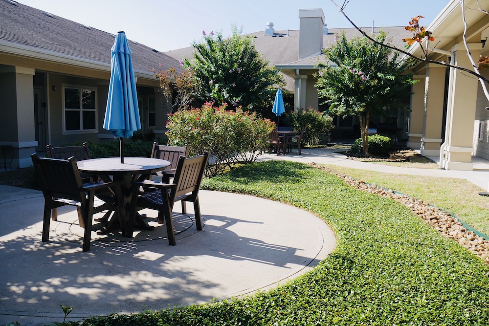 A sunny garden patio with tables, chairs, and umbrellas surrounded by greenery.