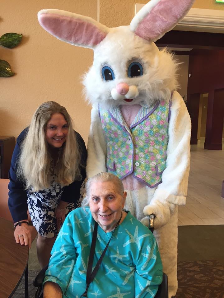 An elderly woman smiles with a person in an Easter Bunny costume, along with another smiling individual indoors.