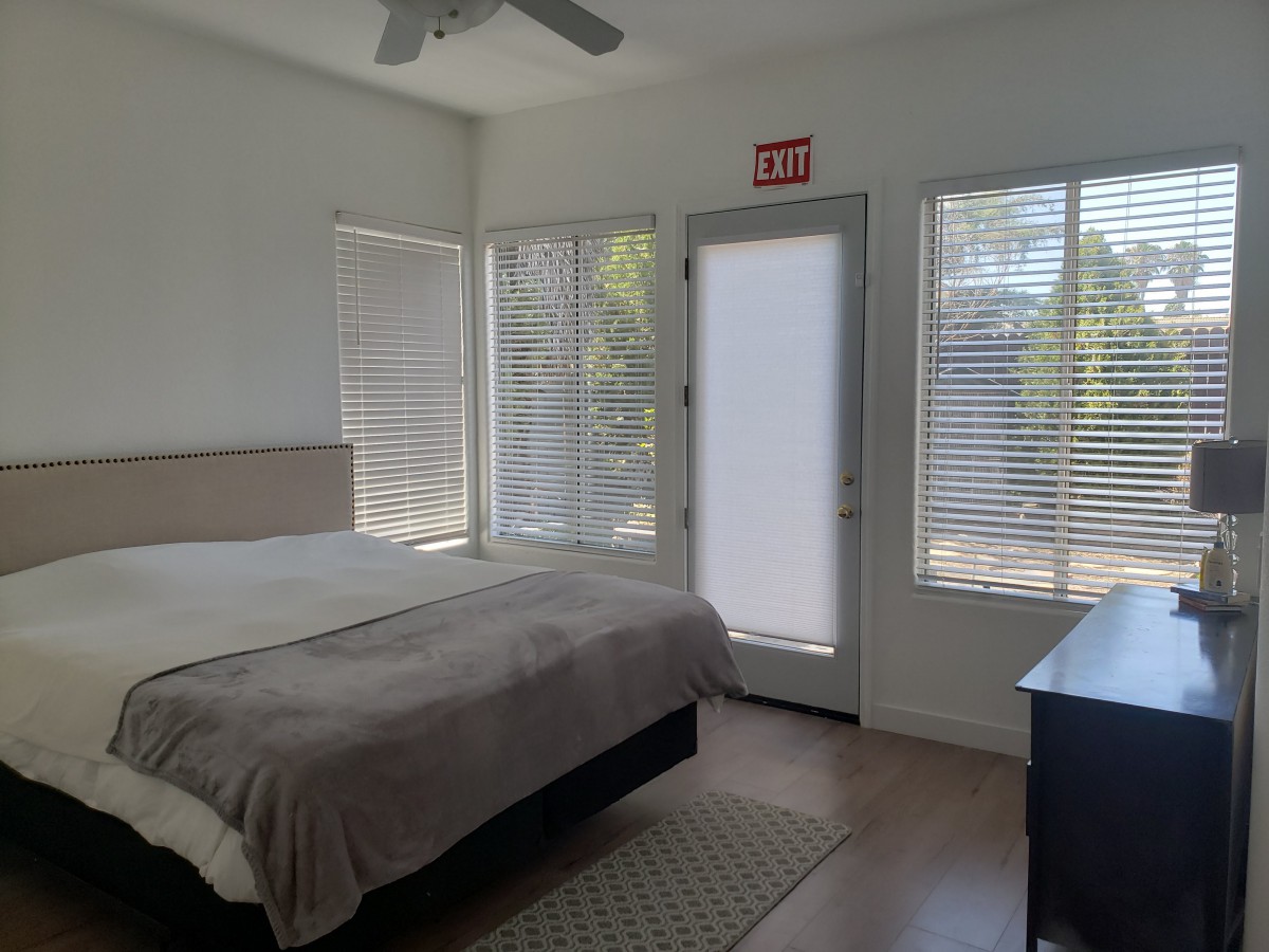 A minimalist bedroom with a bed, dresser, and large windows with blinds.
