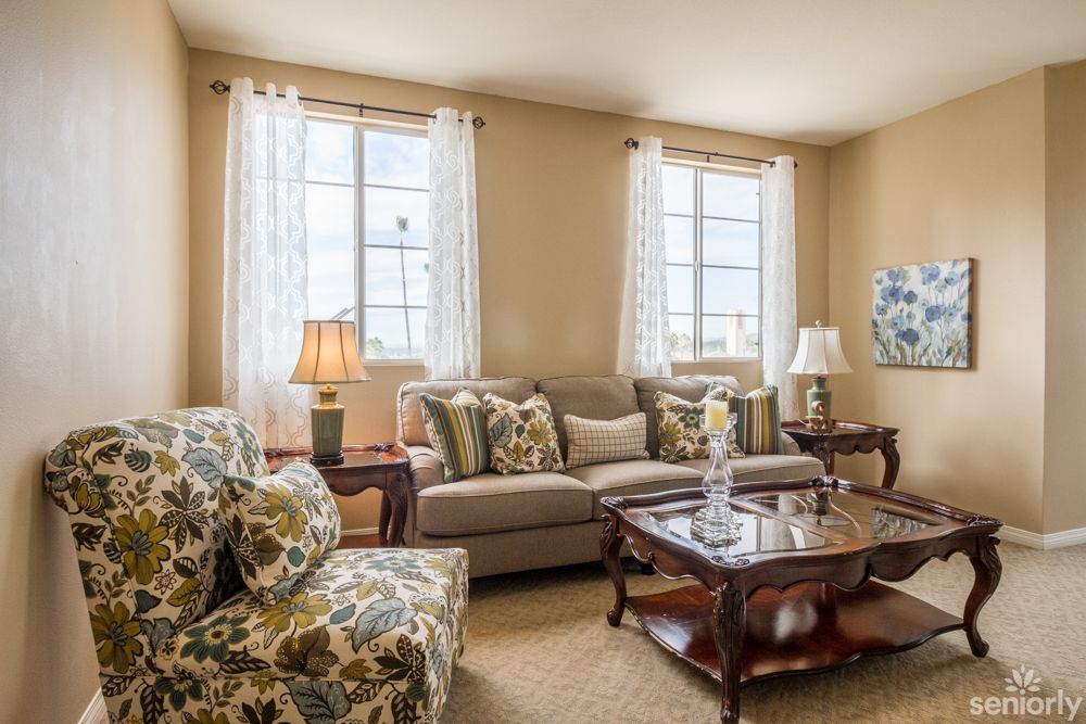A cozy living room featuring a comfortable sofa, armchairs with floral patterns, and a wooden coffee table, complemented by large windows with sheer curtains.