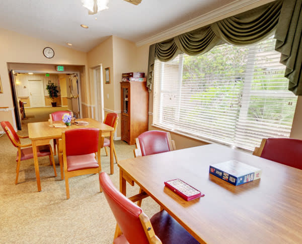A cozy dining area with wooden tables and chairs, large windows with blinds, and carpeted flooring.