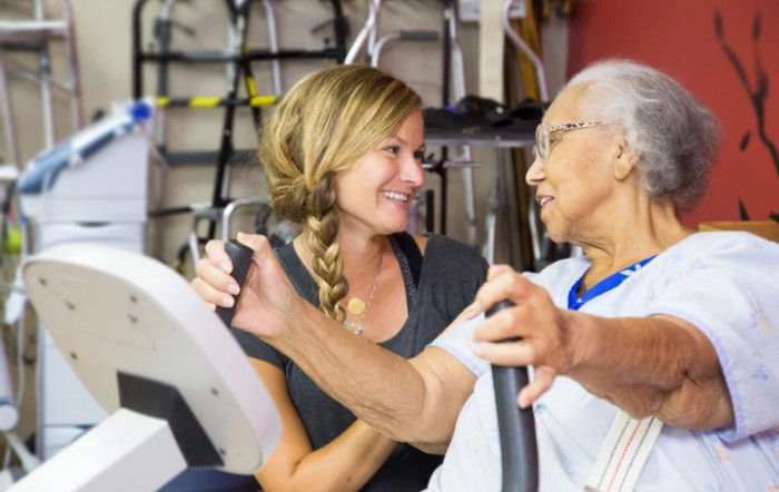 A caregiver assisting an elderly person on exercise equipment.
