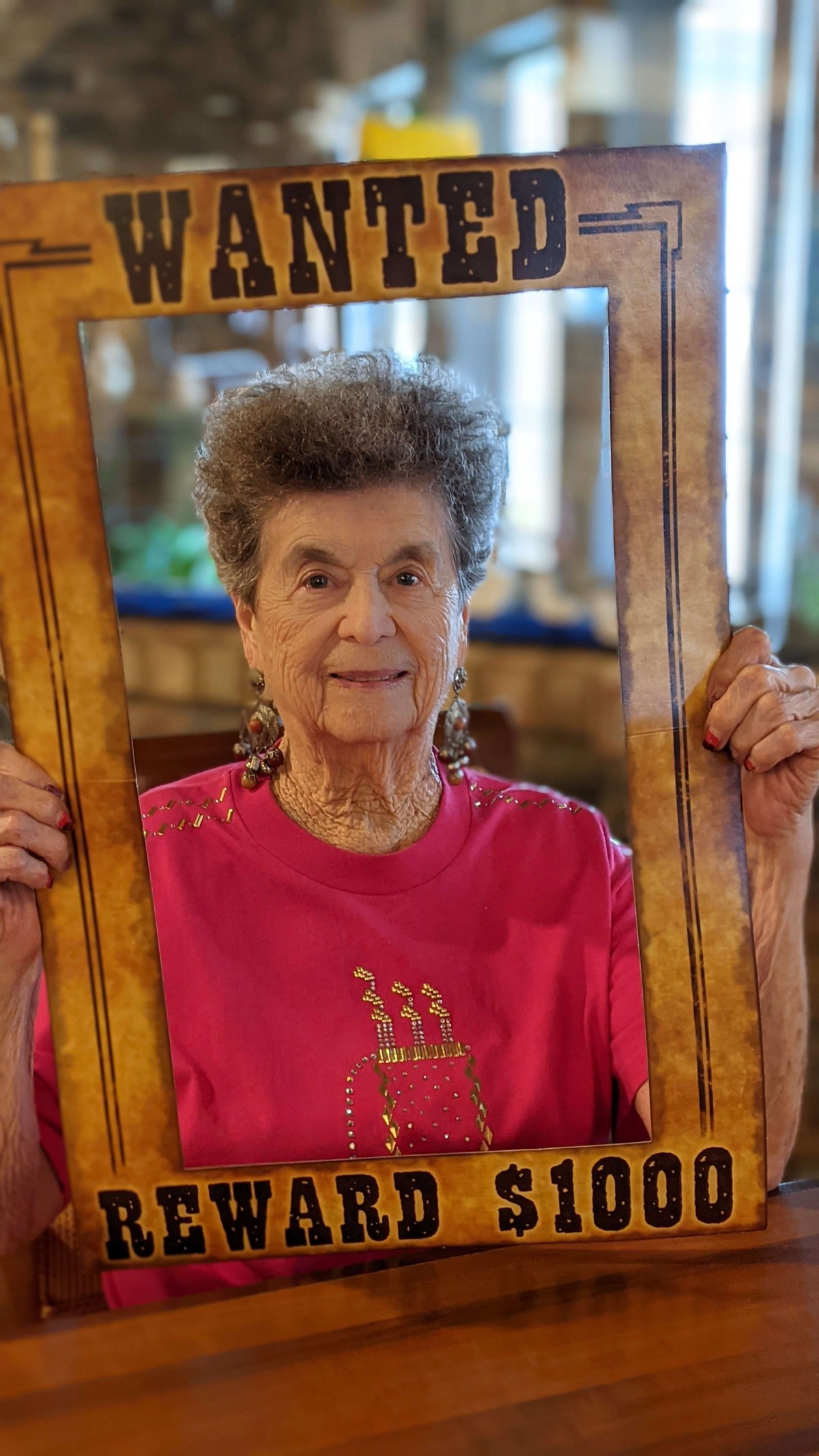 An elderly woman wearing a pink shirt is holding a wooden frame indoors.