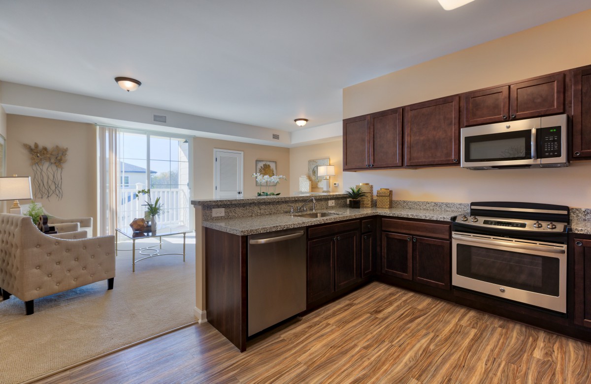A modern kitchen with wooden cabinets and stainless steel appliances, featuring an open view into a living area.
