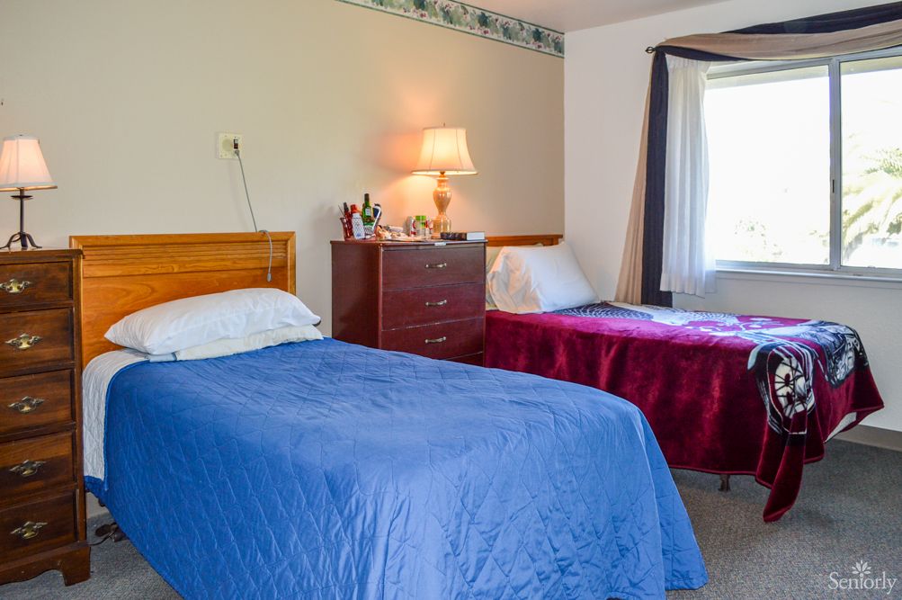 A cozy bedroom with two beds, featuring blue and red bedspreads, a nightstand with a lamp, and a window for natural light.