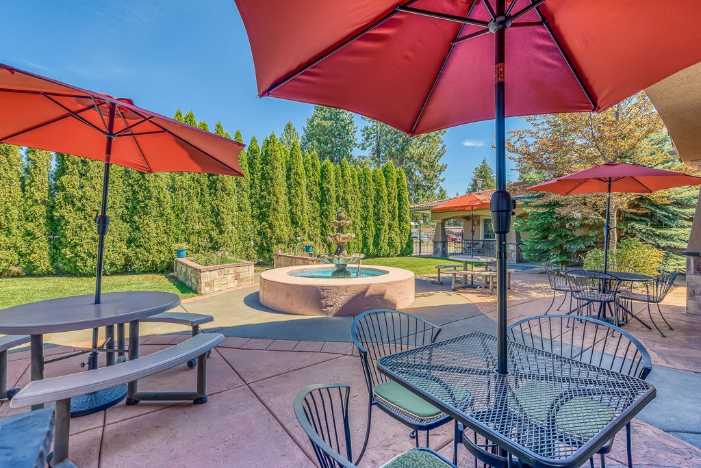 A sunny patio with outdoor seating, featuring tables, chairs, umbrellas, and a central fountain surrounded by lush greenery.