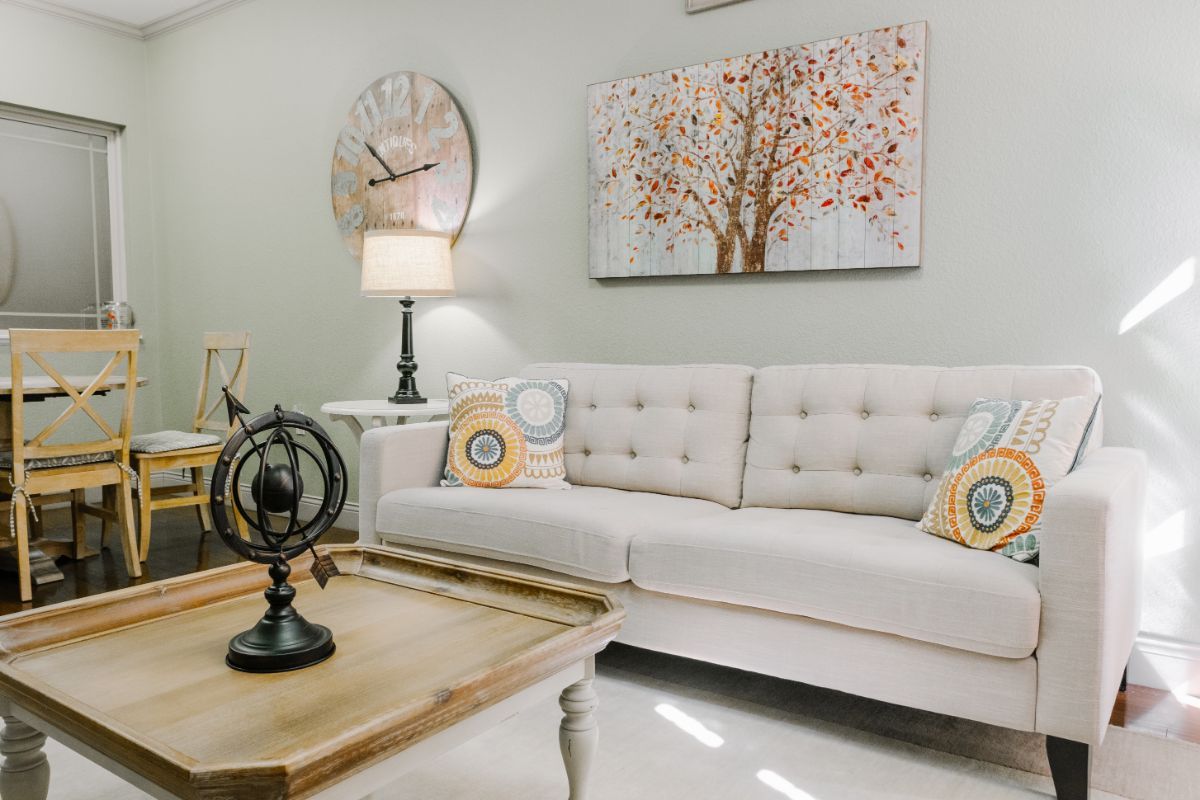 A cozy living room with a plush sofa, adorned with colorful pillows, a wooden coffee table, and decorative elements like a large wall clock and tree art.