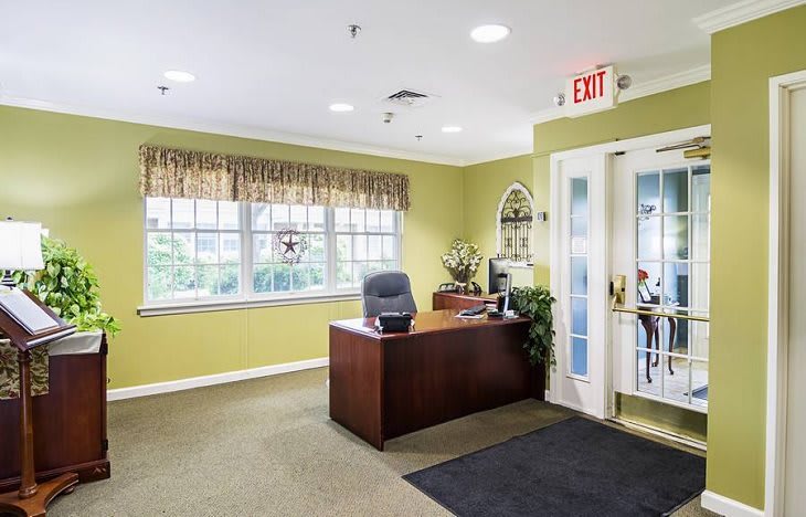 A welcoming reception area with a wooden desk and large windows, accented by plants and an exit sign.