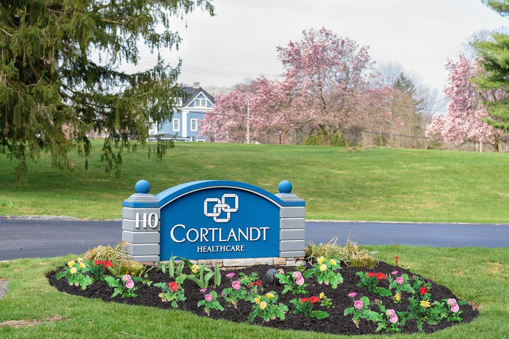 A scenic view of the Cortlandt Hospital sign surrounded by flowers with a building and blooming trees in the background.