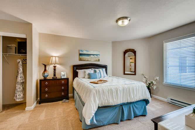 A cozy bedroom with a neatly made bed, adorned with decorative pillows and a blue bed skirt. The room features a wooden nightstand with a lamp, a wall mirror, and a landscape painting above the bed.