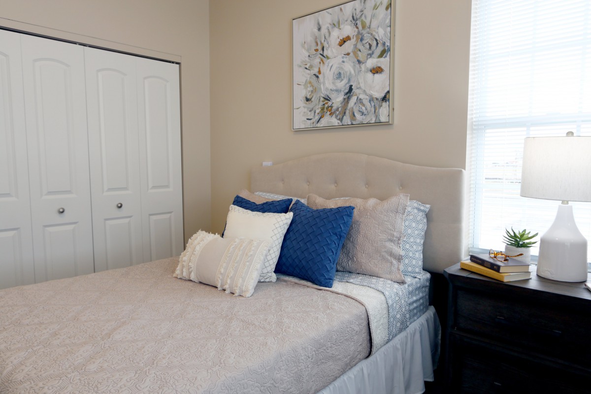 A cozy bedroom featuring a neatly made bed with white and blue pillows, adorned by a floral painting above and natural light streaming through the window.