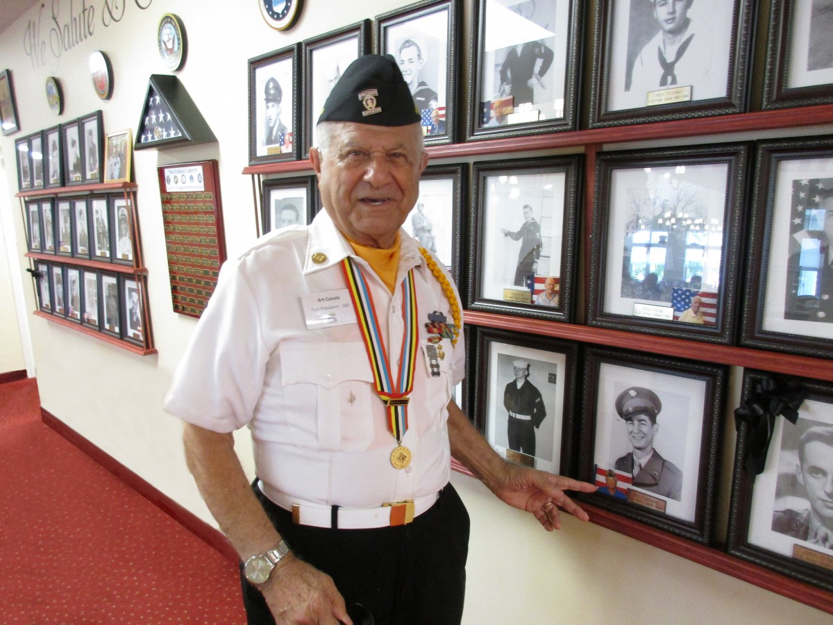 A veteran in uniform standing in front of a wall displaying photographs and medals.