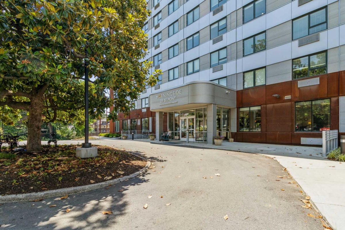 The image shows the entrance of a modern apartment building surrounded by trees.
