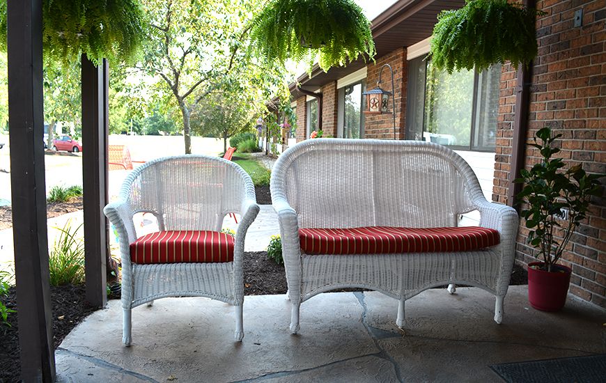A wicker chair and loveseat with red cushions on a patio surrounded by greenery.