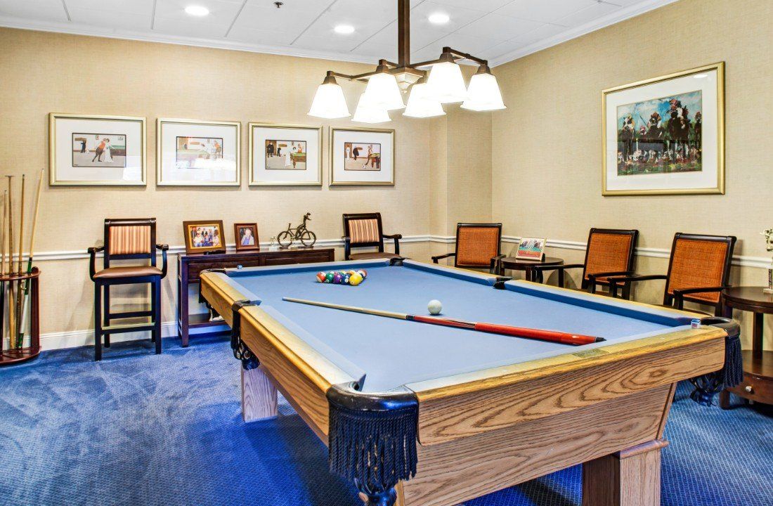 A well-lit game room featuring a pool table with blue felt and surrounding chairs.
