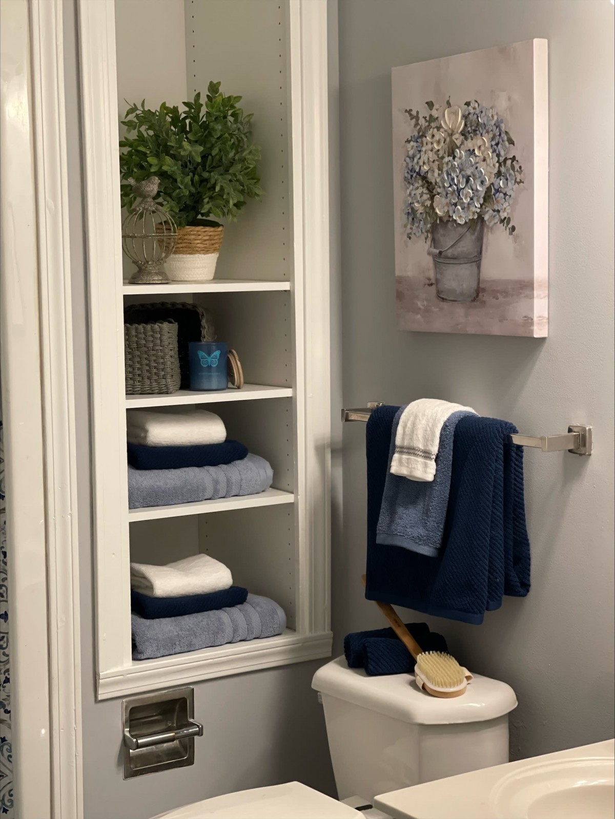 A neatly organized bathroom with blue and white towels, a potted plant on a shelf, and floral artwork on the wall.