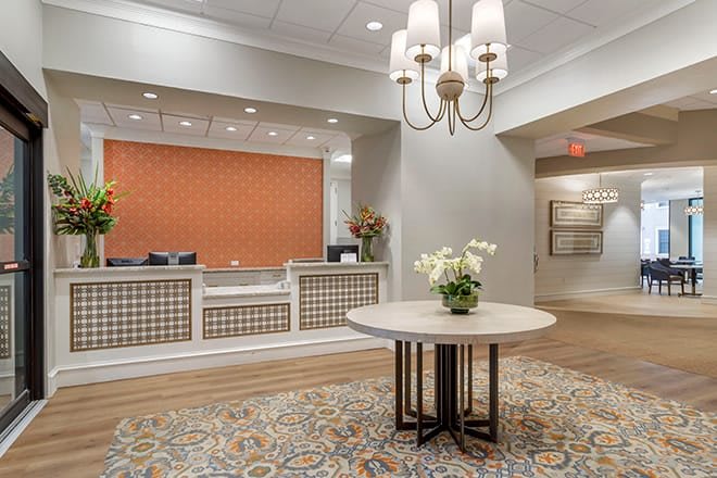 A welcoming hotel lobby with a central table, floral decorations, and a well-lit reception desk.