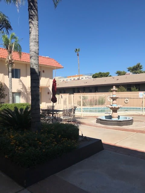 A sunny courtyard of a hotel with a fountain and swimming pool surrounded by patio furniture and palm trees.