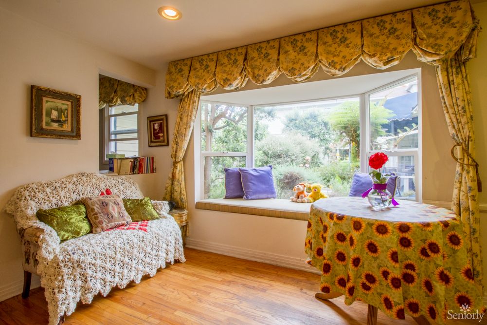 A cozy living room with a window seat overlooking a garden, featuring floral decorations and bright cushions.
