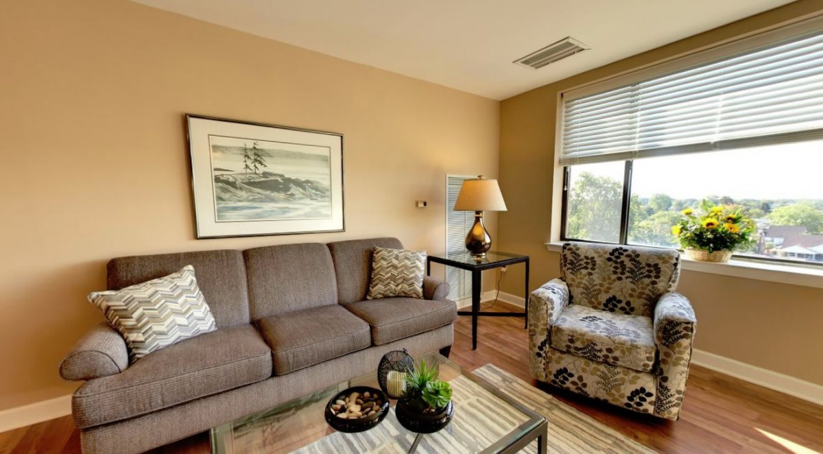 A cozy living room with a sofa, armchair, and coffee table, adorned with artwork and a lamp.