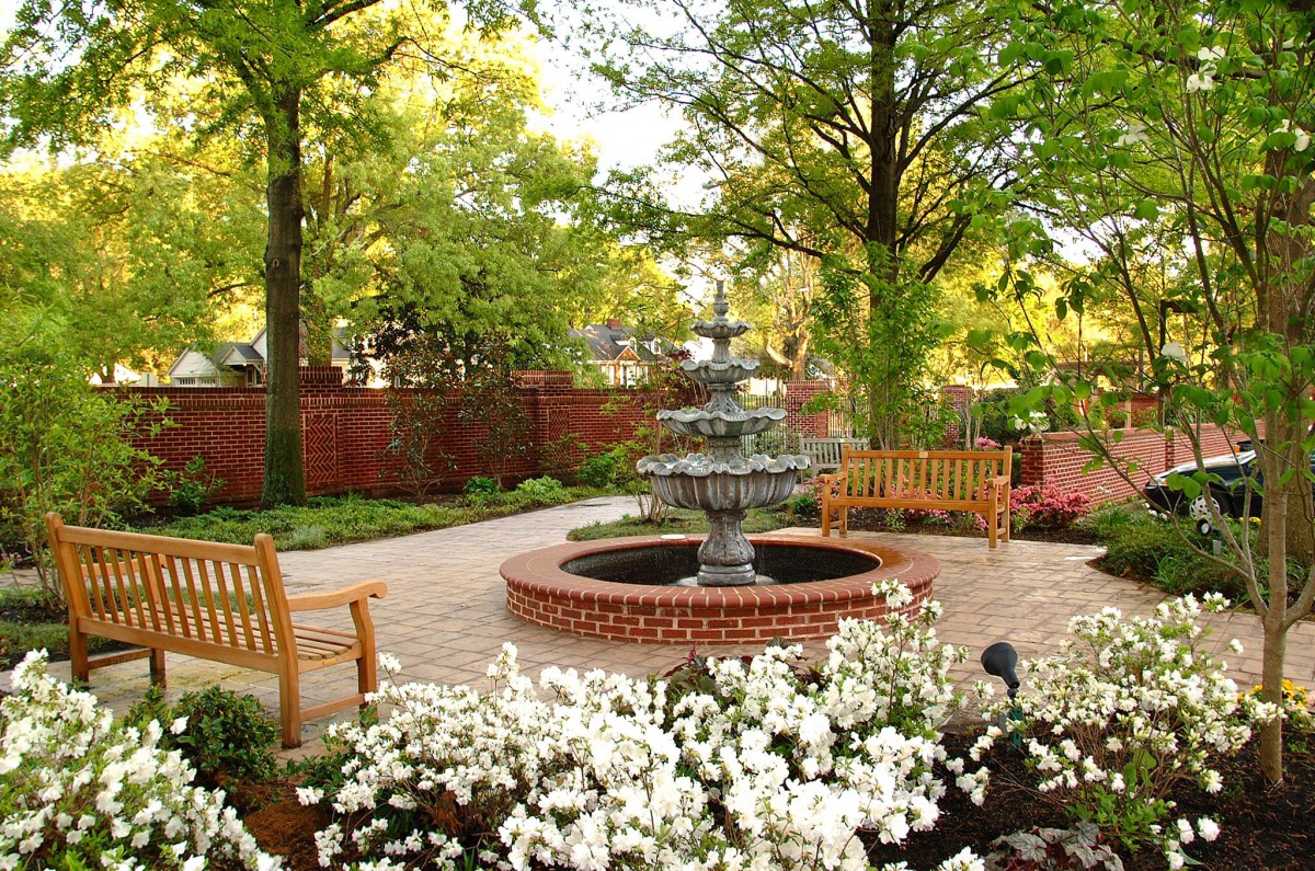 A serene garden with a central fountain surrounded by benches, trees, and blooming flowers.