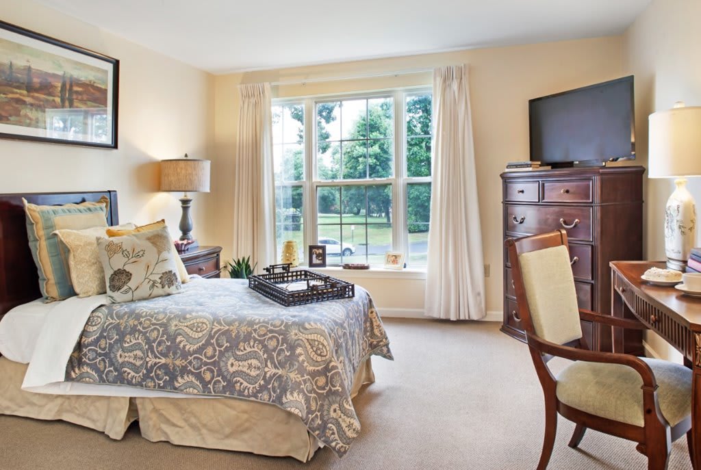 A cozy bedroom with a large window, furnished with a bed, bedside lamp, dresser, and television.